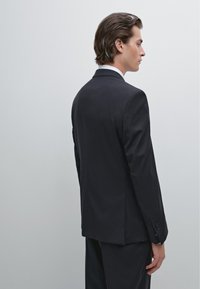Sort jakkesætjakke lavet af glat stof, med enkelt slids bagpå og et slankt, skræddersyet fit. Hvid skjorte synlig ved kraven.