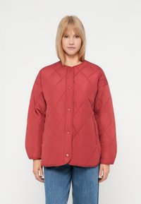 Kvinne iført en rød, quiltet knappjakke med lange ermer og blå jeans, som står foran en enkel hvit bakgrunn.