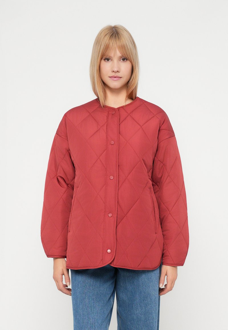 Kvinne iført en rød, quiltet knappjakke med lange ermer og blå jeans, som står foran en enkel hvit bakgrunn.