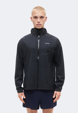 Mladý muž s krátkymi vlasmi, ktorý má na sebe čiernu Salomon bundu na zips a námornícke modré kraťasy, stojaci pred jednoduchým svetlým pozadím.