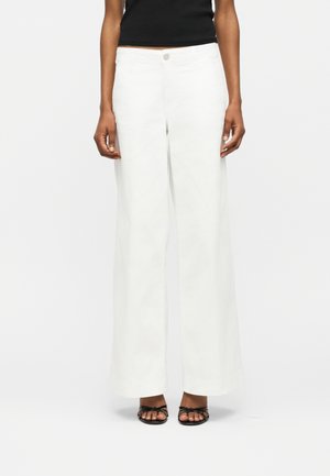 Άτομο που φοράει λευκό παντελόνι με ψηλή μέση και φαρδιά μπάσκα, μαύρο αμάνικο τοπ και μαύρα σανδάλια με λουράκια και ανοιχτά δάχτυλα, στέκεται σε λευκό φόντο.