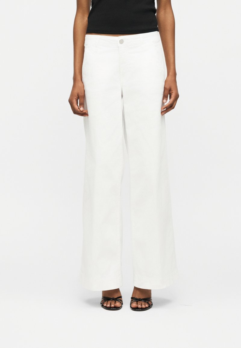 Άτομο που φοράει λευκό παντελόνι με ψηλή μέση και φαρδιά μπάσκα, μαύρο αμάνικο τοπ και μαύρα σανδάλια με λουράκια και ανοιχτά δάχτυλα, στέκεται σε λευκό φόντο.