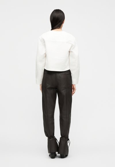 Γυναίκα με μακριά σκούρα μαλλιά που φοράει λευκό κοντό μπουφάν, μαύρο δερμάτινο ψηλόμεσο παντελόνι και μαύρα μποτάκια με τακούνι, στέκεται με την πλάτη προς τα έξω.