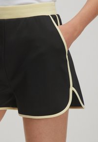 Shorts de sport noirs avec une taille et des bordures contrastantes de couleur jaune clair, dotés de poches latérales et d'un design d'ourlet incurvé.