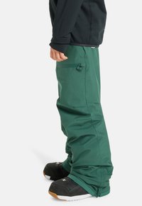 Groene waterdichte skibroek met een losse pasvorm, voorzien van ritszakken en een verstelbare boei, gecombineerd met zwarte sneeuwlaarzen.