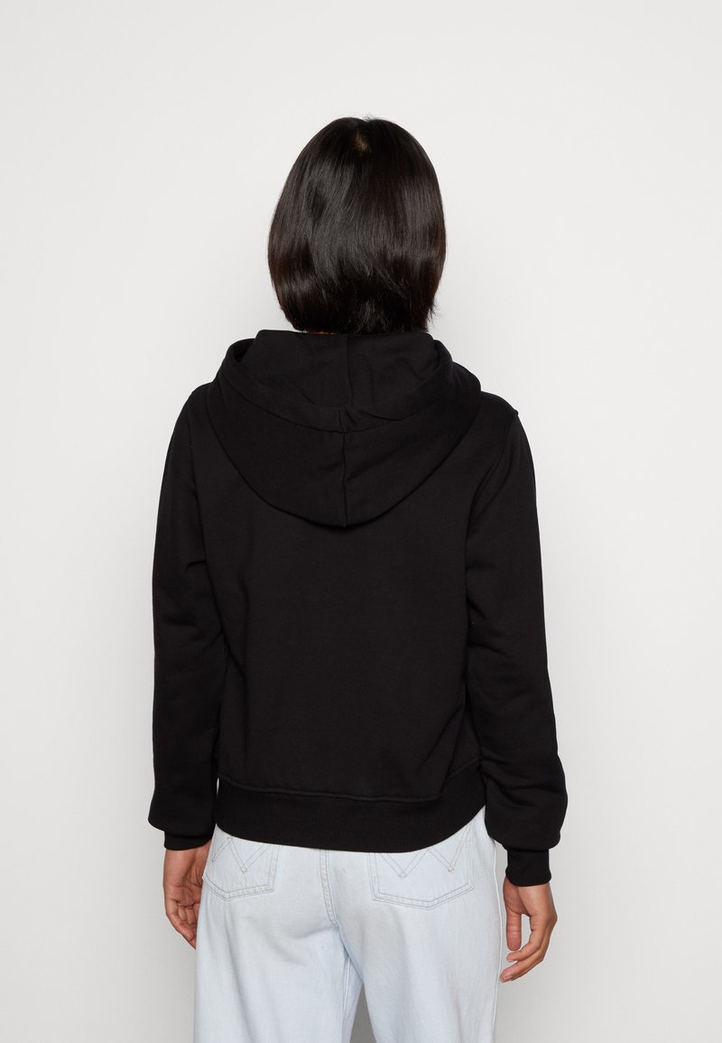 Schwarzer Hoodie mit lockerer Passform, ausgestattet mit einer großen Kapuze, gerippten Bündchen und einem Saum. Sichtbare Nähte und ein schlichtes Design, getragen über einer hellen Jeans.