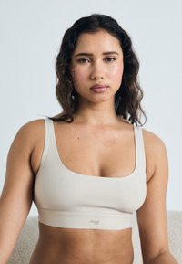 Beige bralette med firkantet halsutskjæring, laget av mykt materiale, med en bred underbånd med logo-detall. Jevn tekstur og minimalistisk design.