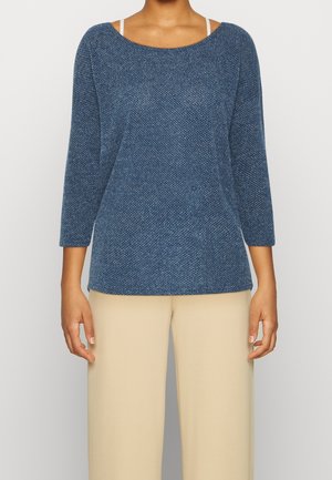 Vrouw die een blauw textuurdriekwarts mouwtop draagt met beige wijde broekspijpen tegen een effen achtergrond.