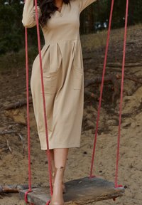 Femme en robe beige et bottines montant à la cheville, debout sur une balançoire en bois avec des cordes rouges dans un cadre extérieur sablonneux avec des arbres.
