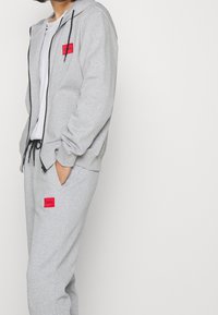 Sudadera gris con cremallera y cordones, con un parche de logo rojo. Combinada con pantalones deportivos grises a juego y un parche de logo rojo adicional.