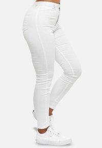 Mujer vestida con jeans skinny blancos y zapatillas blancas con cordones, de pie con una rodilla ligeramente doblada.