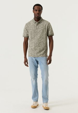 Mann steht vor neutralem Hintergrund, trägt ein hellgrünes Poloshirt mit Blumenmuster, hellblaue Jeans und beige Turnschuhe.