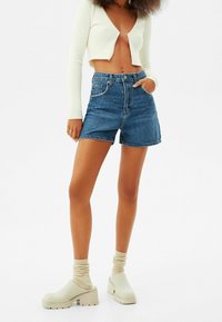 Szorty dżinsowe w średnim odcieniu niebieskiego, z wysokim stanem, prostym krojem i postrzępionym brzegiem; zestawione z białym, krótkim prążkowanym topem i beżowymi chodakami.