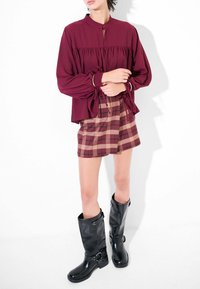 Blusa lunga bordeaux con davanti arricciato e dettagli con nodo, abbinata a una gonna mini scozzese nei toni beige e bordeaux, e stivali neri al ginocchio.