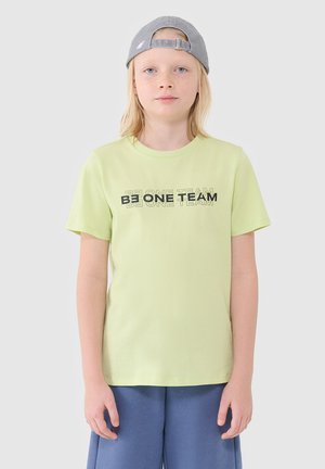 Jeune personne aux cheveux clairs portant une casquette grise à l'envers, un t-shirt vert clair avec le texte "BE ONE TEAM" et un short bleu, debout devant un fond uni.