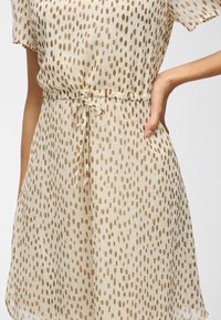 Femme portant une robe beige clair à manches courtes avec un motif de points ovales marron, nouée à la taille avec une fine ceinture en tissu.