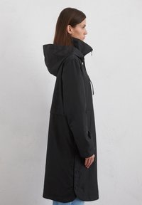 Manteau noir à capuche en tissu lisse et imperméable, doté d'un col haut et de poches latérales. Boutons le long de l'ourlet pour un ajustement réglable.