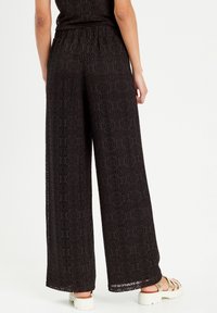 Schwarze Spitzen-Hosen mit weiter Passform und strukturiertem Muster, elastischem Bund und detaillierter Stickerei. Kombiniert mit hellen Sandalen.
