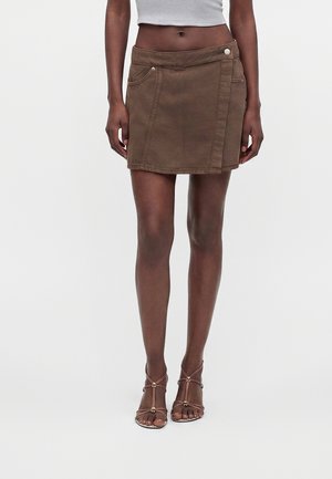 Femme portant une jupe portefeuille marron avec des poches, un top court blanc et des talons marron à brides et bouts ouverts sur un fond uni.
