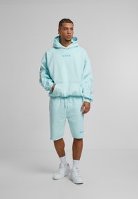 Lyseblå hettegenser og shorts sett. Hettegenseren har en forkjøpslomme og dekorativt mønster på ermene. Fullført med hvite joggesko og sokker.