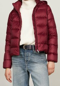 Bordowa pikowana kurtka z wysokim kołnierzem, zamkiem błyskawicznym i odpinanym kapturem; posiada pikowaną fakturę i elastyczne mankiety. W zestawie z białym swetrem i niebieskimi dżinsami.