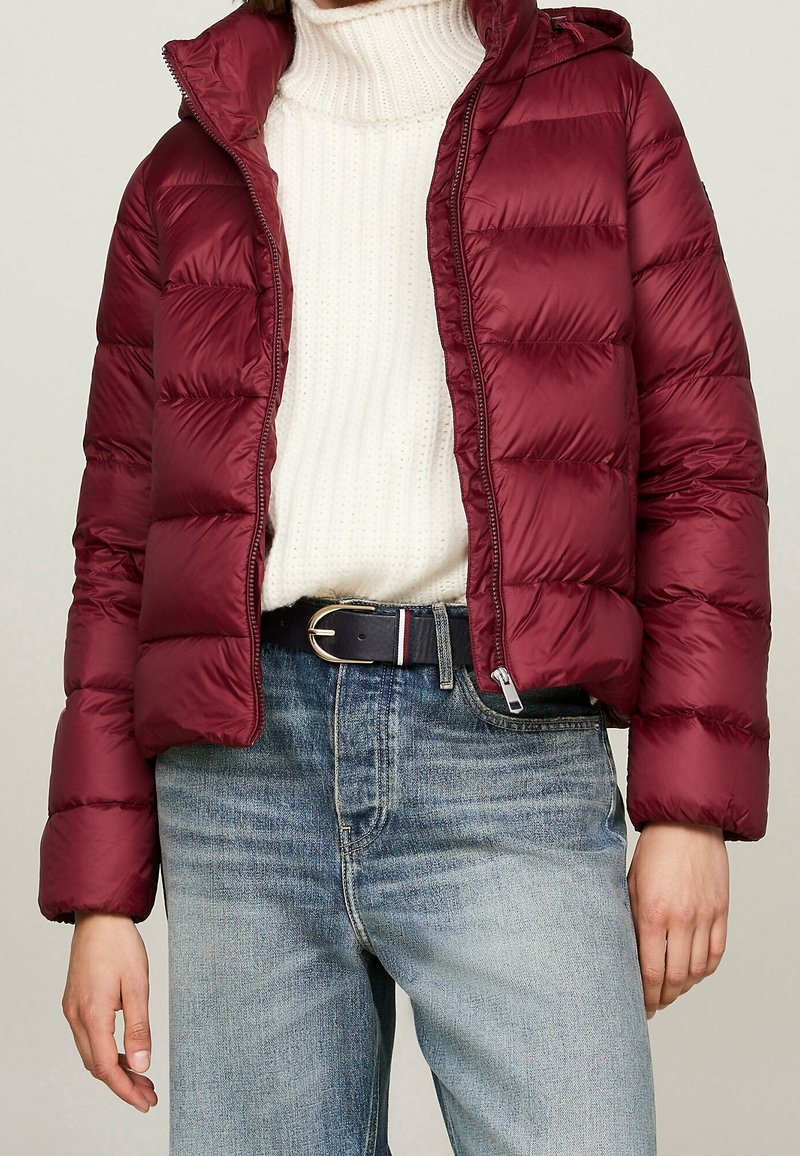 Bordowa pikowana kurtka z wysokim kołnierzem, zamkiem błyskawicznym i odpinanym kapturem; posiada pikowaną fakturę i elastyczne mankiety. W zestawie z białym swetrem i niebieskimi dżinsami.