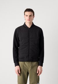 Veste bomber noire avec un devant matelassé, des poignets et un col côtelés. Accompagnée d'un pantalon vert olive. Texture de tissu lisse.