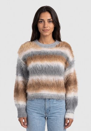 Donna con capelli lunghi e scuri che indossa un maglione a righe morbido beige, grigio e bianco e jeans azzurri chiari, in piedi davanti a uno sfondo semplice.