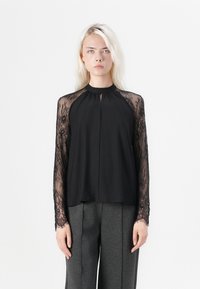 Black top with long sheer lace sleeves, rounded neckline, and pleated front design, paired with gray trousers. Smooth fabric texture.