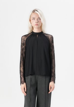 Svart topp med lange gjennomsiktige blondesleeves, rund halslinje og plissert frontdesign, kombinert med grå bukser. Glatt stofftekstur.