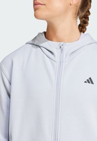 Harmaa vetoketjuhuppari, joka on valmistettu pehmeästä kankaasta ja siinä on nyörillä säädettävä huppu sekä pieni musta logo vasemmassa rintakohdassa.