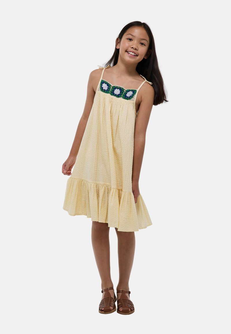 Robe sans manches jaune en tissu texturé, avec un ourlet à volants et des accents floraux en crochet bleus et verts autour de l'encolure.