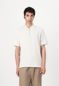 White short-sleeve polo shirt with a zippered neck, made of textured fabric. Featured on a model with beige trousers and arm tattoo.