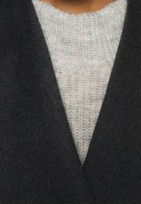 Schwarzer Wollmantel mit einer weichen Textur, ausgestattet mit einem breiten Kragen, über einem hellgrauen Strickpullover mit einem gerippten Muster getragen.