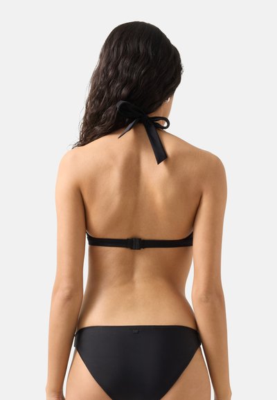 Kvinde med langt mørkt hår bundet med en sløjfe iført sort halterbikinitop og matchende bikinitrusser, vist bagfra.