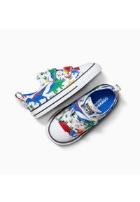 Sneakers per bambini con stampa di dinosauri in blu, verde e rosso su tela bianca. Presentano fibbie in velcro e una suola in gomma.