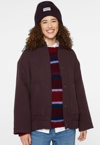 Dunkelviolet, geknöpfte Jacke mit zwei vorderen Taschen, strukturiertem Stoff. Darunter ein mehrfarbig gestreifter Pullover und eine passende Mütze.