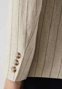 Beige textured fabric with narrow black stripes, featuring three round light brown buttons on the cuff of a sleeve.