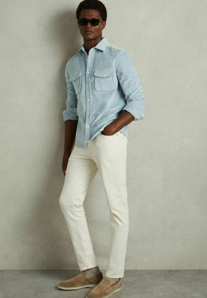 Light blue, long-sleeve, button-up shirt with chest pockets, paired with white fitted pants and tan slip-on shoes. Sunglasses worn.