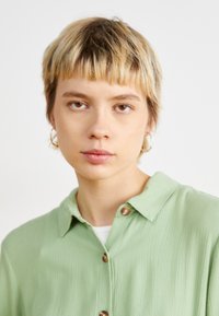 Chemise boutonnée légère verte avec un col, en tissu texturé et ornée de boutons marron proéminents. Le modèle a les cheveux blonds courts.