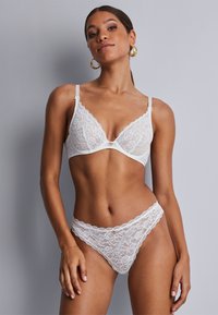 Ensemble de lingerie en dentelle blanche comprenant un soutien-gorge triangulaire avec des bords festonnés et un accent décoratif doré, accompagné d'un string en dentelle assorti.