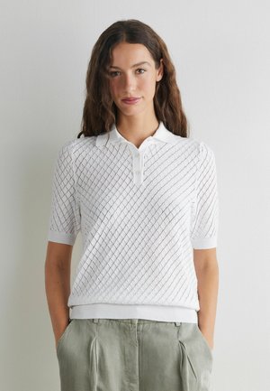 Kvinde med langt, bølget brunt hår iført en hvid kortærmet strikket polotrøje og lysegrå bukser, stående mod en ensfarvet hvid væg.