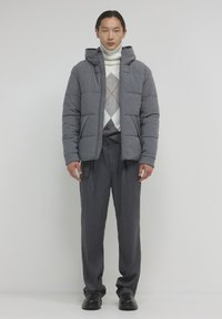 Giubbotto puffer grigio con cappuccio, design trapuntato, chiusura frontale con zip, indossato sopra un maglione a fantasia e pantaloni grigi, completato con scarpe nere.
