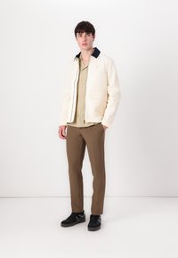 Off-white lightweight jacket with navy collar, two front pockets; paired with a green striped shirt and brown trousers, completing the outfit.