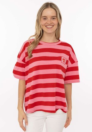 Lächelnde Frau trägt ein locker geschnittenes rot-rosa gestreiftes T-Shirt mit kleinem Brustlogo und weiße Hose, steht vor einem weißen Hintergrund.