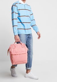 Světle modré polo triko s dlouhým rukávem s hnědými pruhy, šedé džíny, bílé tenisky a růžový batoh s předním zipem a dvojitými uchy.