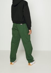 Černá mikina s kapucí v kombinaci se zelenými cargo kalhotami. Kalhoty mají boční kapsy a nášivku. Boty jsou bílé s černým hvězdným akcentem.