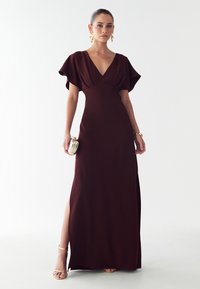 Burgundská večerná toaleta so širokými rukávmi, hlbokým výstrihom do V, bočným rozparkom a hladkou textúrou. Doplnkovaná kovovou kabelkou a sandálmi na tenkých podpätkoch.