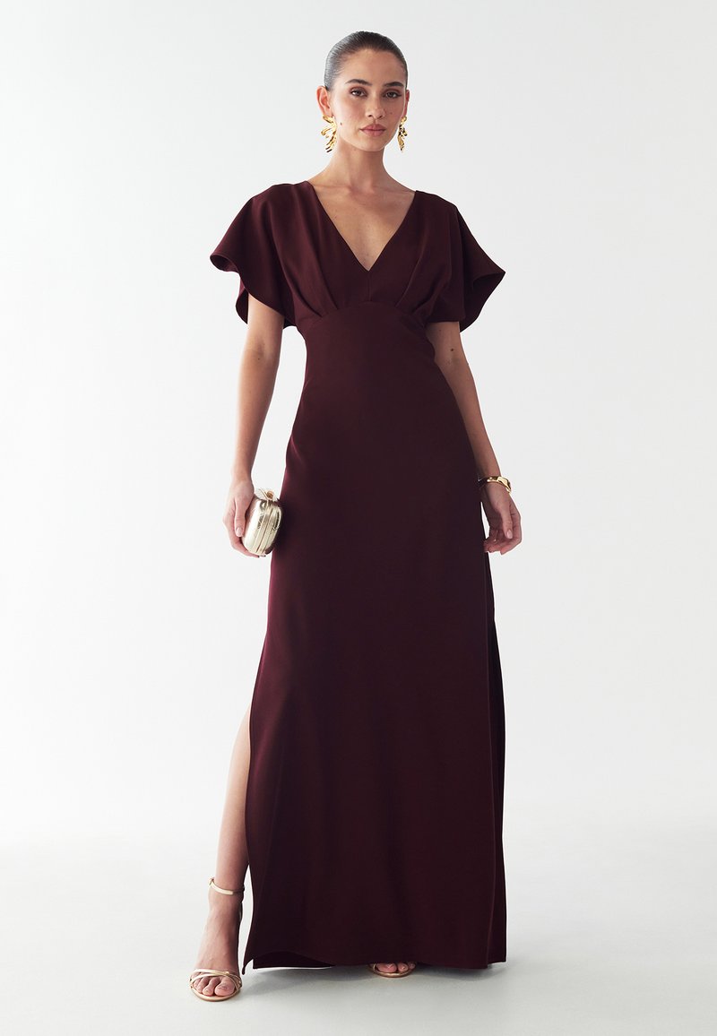 Burgundská večerná toaleta so širokými rukávmi, hlbokým výstrihom do V, bočným rozparkom a hladkou textúrou. Doplnkovaná kovovou kabelkou a sandálmi na tenkých podpätkoch.