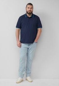 Granatowa koszulka polo z turkusowymi akcentami, w zestawie z jasnoniebieskimi dżinsami i białymi trampkami. Koszulka ma kołnierzyk i krótkie rękawy.